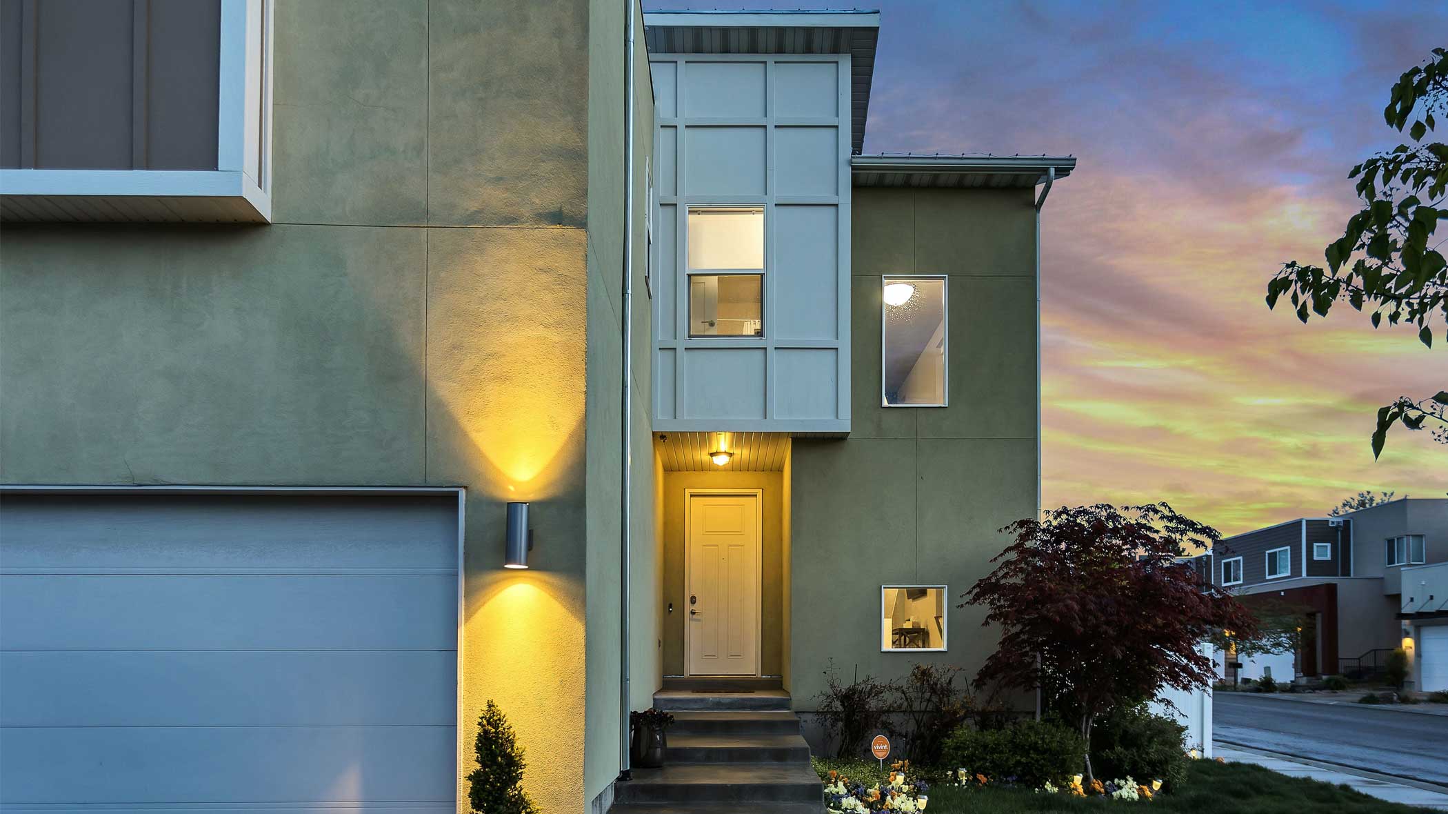 Frontalansicht eines Hauseingangs bei Sonnenuntergang. Das Haus wird von Spots beleuchtet, die neben der Garage und oberhalb der Eingangstür angebracht sind. Der Himmel im Hintergrund ist rosa, während die Sonne gerade untergeht.